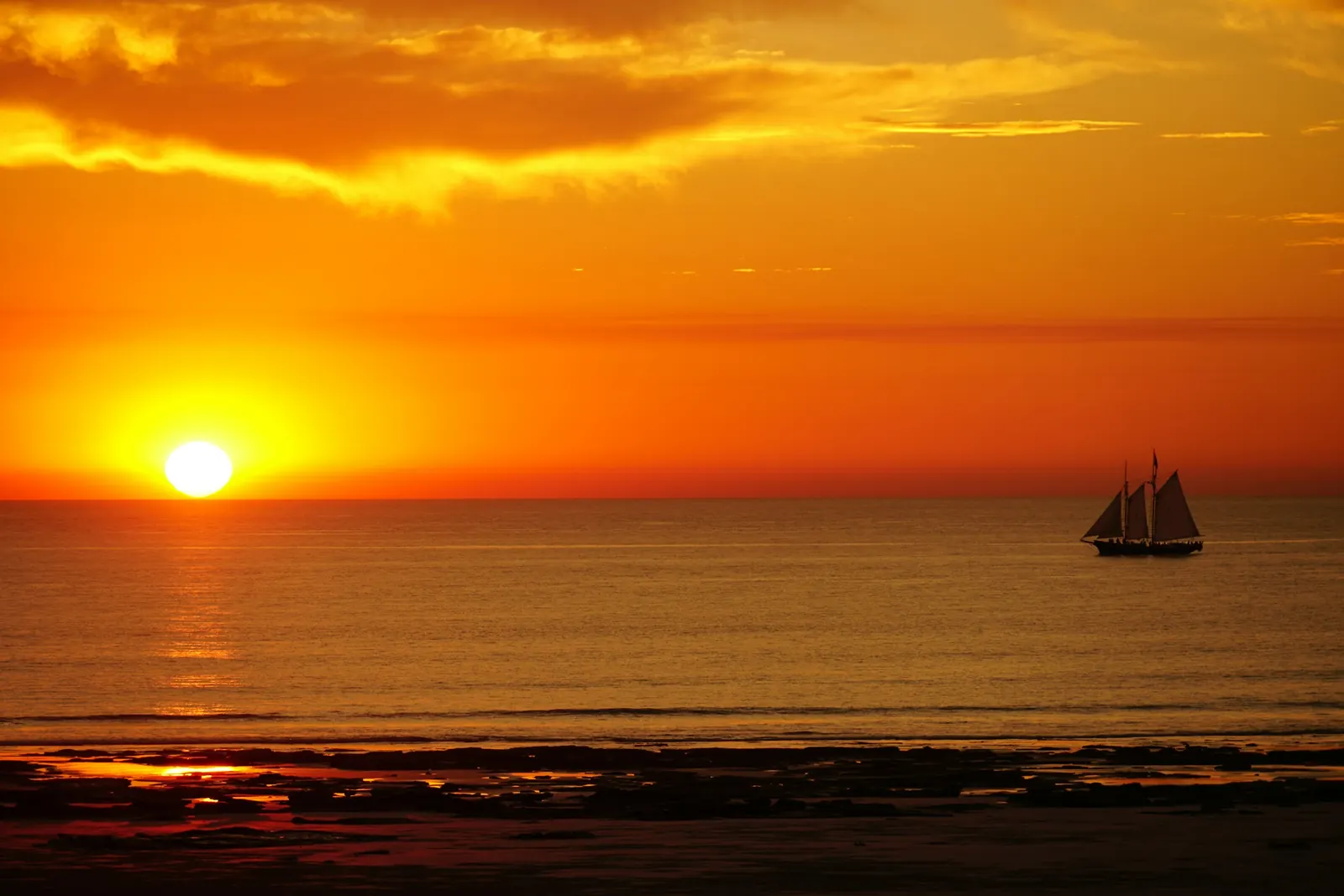 A two-masted sailing ship crosses the horizon at sunset with the low sun on the left, seen from Cable Beach